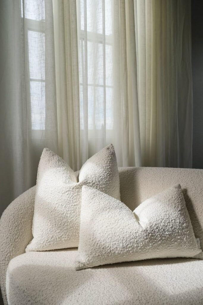 Cozy white pillows on a textured sofa in soft daylight filtering through sheer curtains.