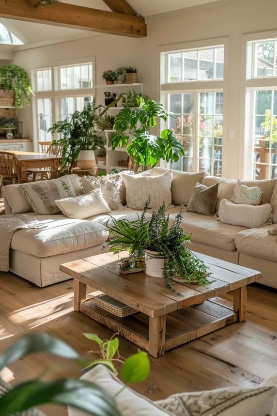 Bright cozy living room with beige sectional sofa, wooden coffee table, and lush green plants by large sunny windows.