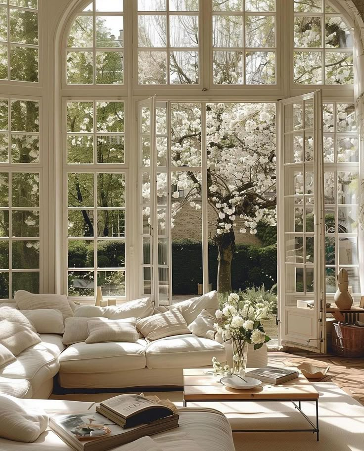 Bright sunlit living room with large windows, white sofa, and blooming trees outside. Cozy and elegant interior.