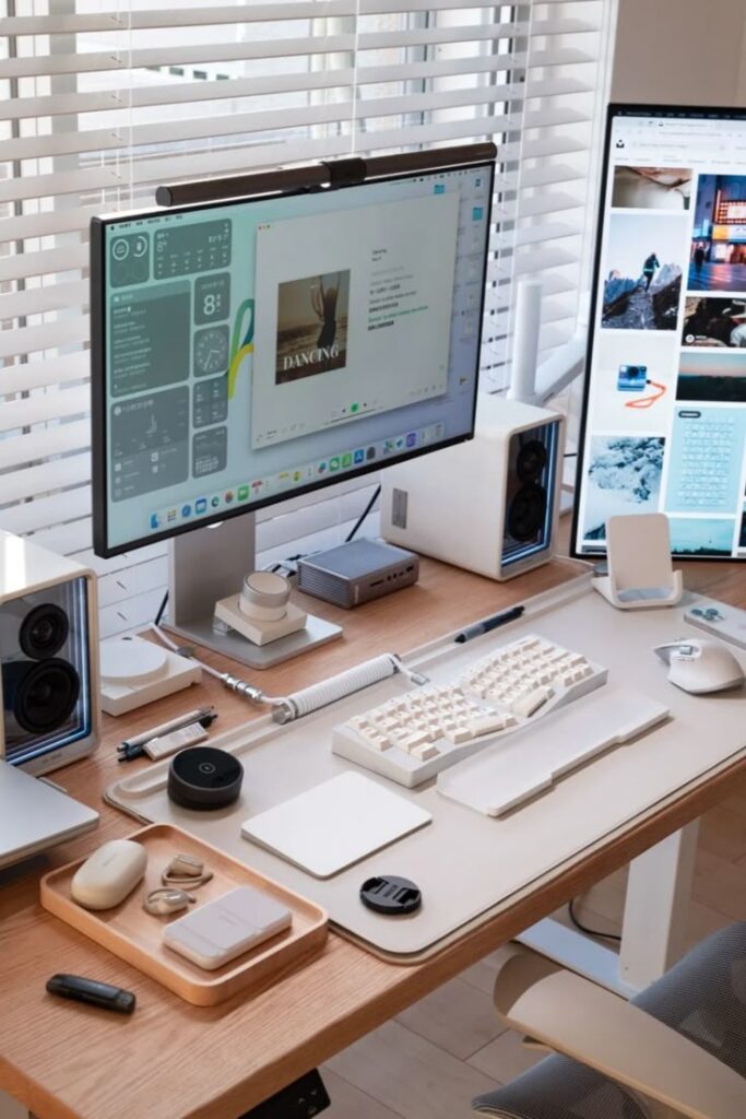 Minimalist home office setup with dual monitors, keyboard, and tech accessories on a wooden desk near window blinds.