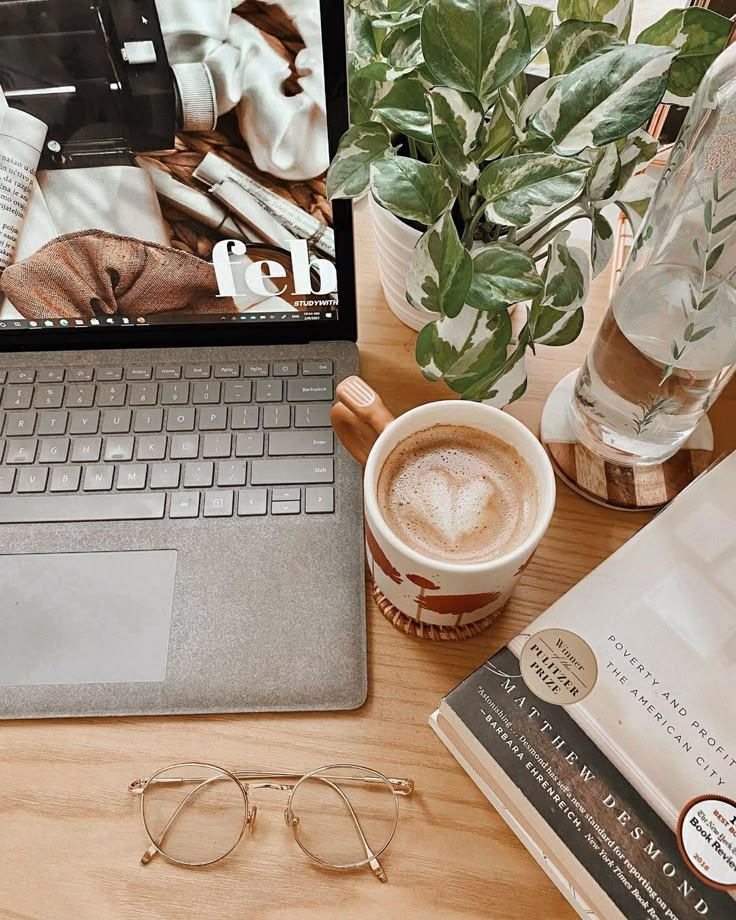 Laptop and coffee on desk with glasses, book, and plant; cozy workspace setup for productivity.