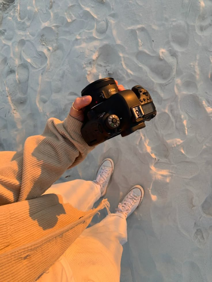 Person holding a camera on a sandy beach, capturing footprints during sunset.