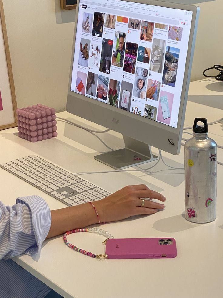 Person browsing Pinterest on a desktop with pink phone and water bottle nearby on a white desk.