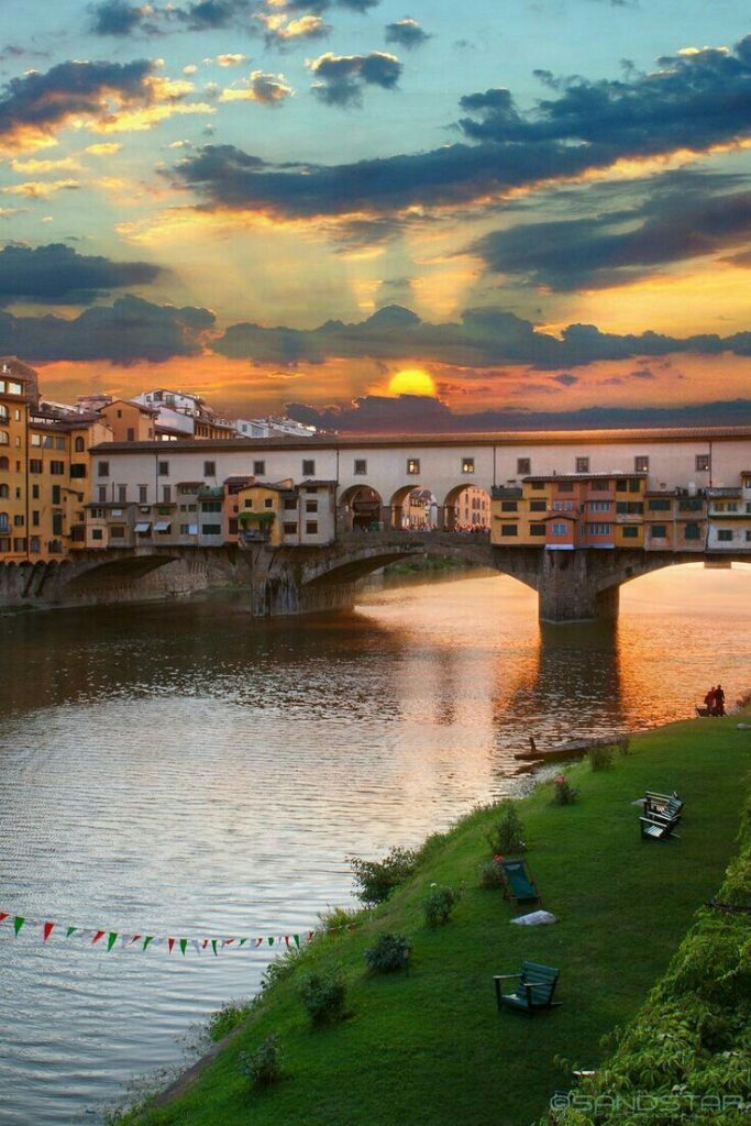 Sunset over Florence's Ponte Vecchio bridge reflecting on the Arno River, with vibrant sky and lush riverside.