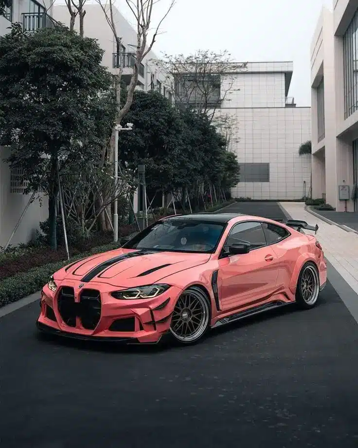 Custom pink sports car with black accents parked on a city street, surrounded by modern buildings and trees.