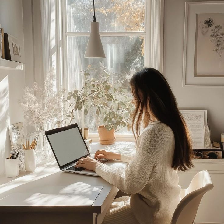 Woman working on laptop at a cozy desk by a sunlit window with plants and decor, creating a tranquil workspace ambiance.