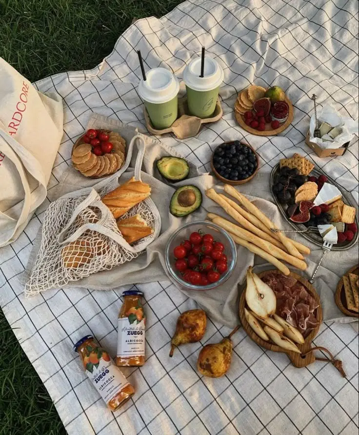 Picnic spread with fruits, bread, juice, and coffee on a checkered blanket outdoors.