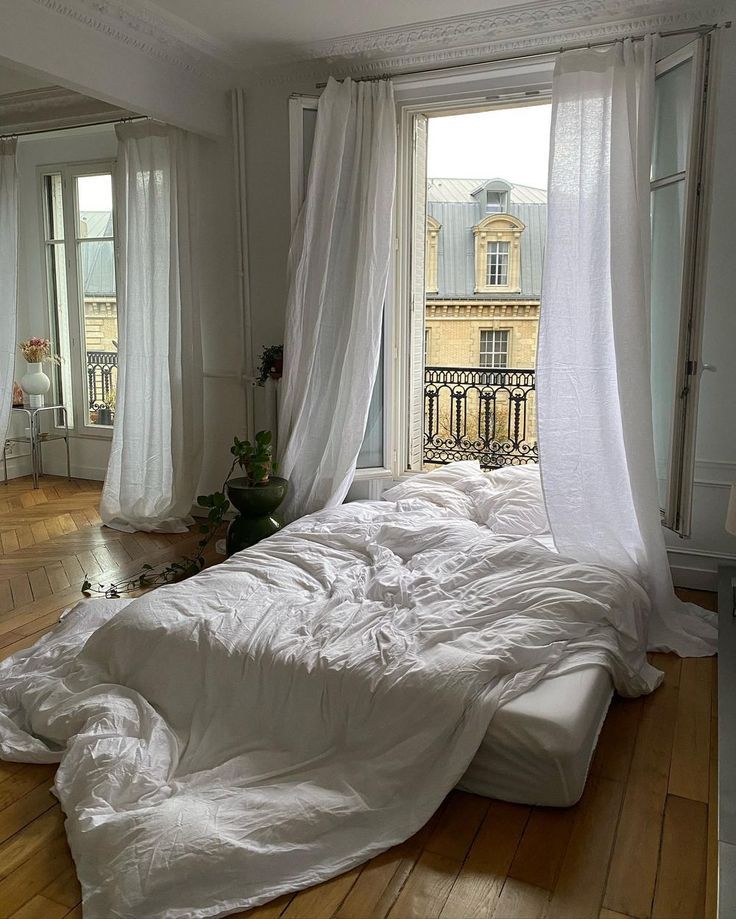 Cozy bedroom with unmade bed, white curtains, and a view of classic architecture through a large window.