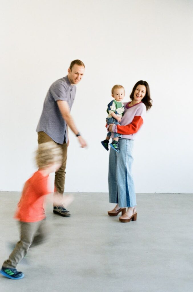 Family enjoying playful moment indoors, parents smiling and holding children in bright, casual outfits.