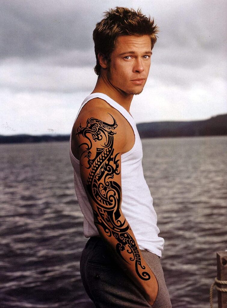 Man in a white tank top with intricate arm tattoo, standing by a lake.