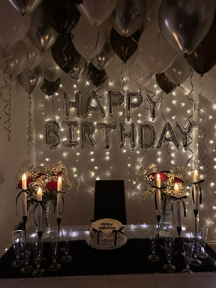 Elegant birthday setup with candles, cake, flowers, and balloons in a dimly lit room with Happy Birthday balloons.