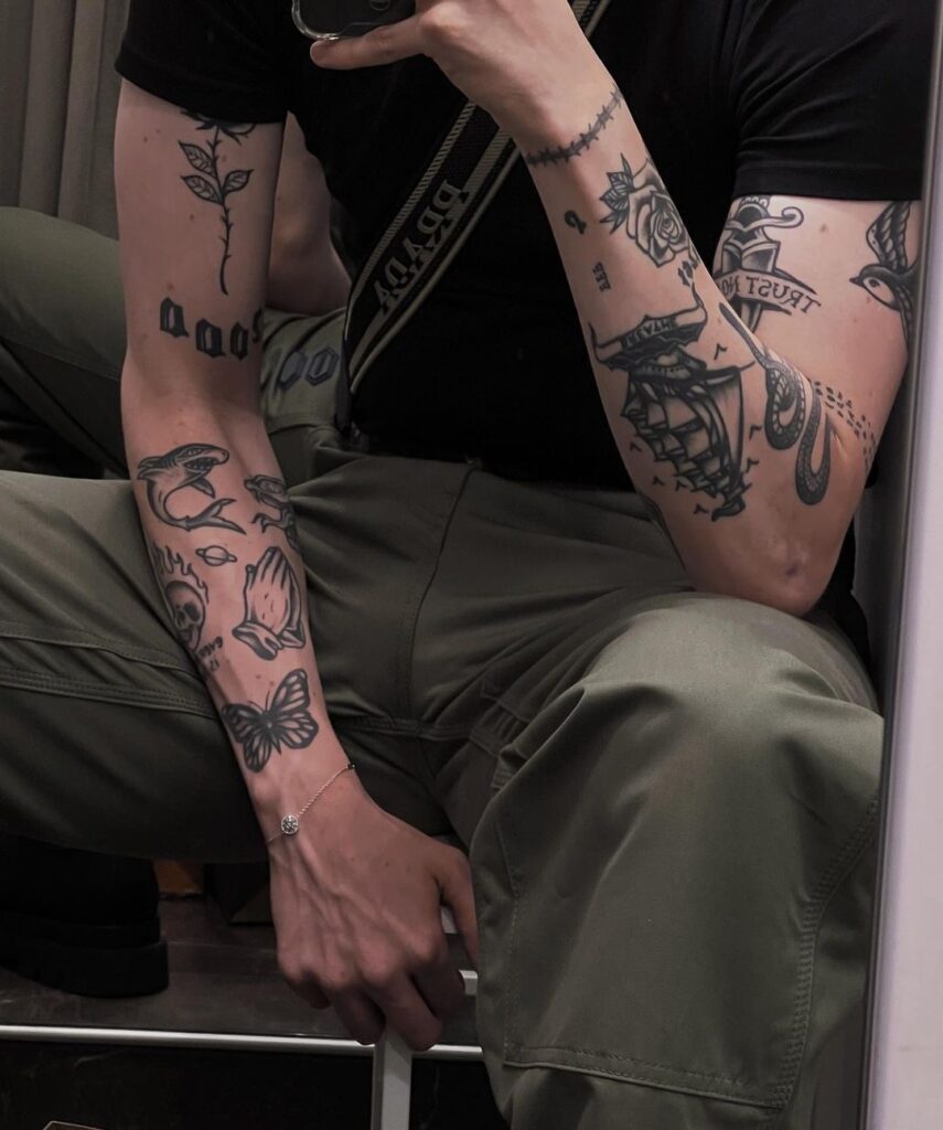 Man with detailed arm tattoos sits in mirror selfie, wearing green pants and a black shirt.