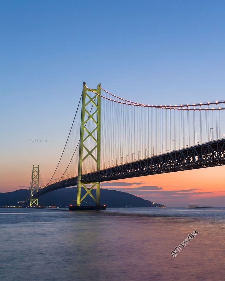 Suspension bridge over calm waters at sunset, vibrant skies enhancing its structure. Ideal travel destination scene.