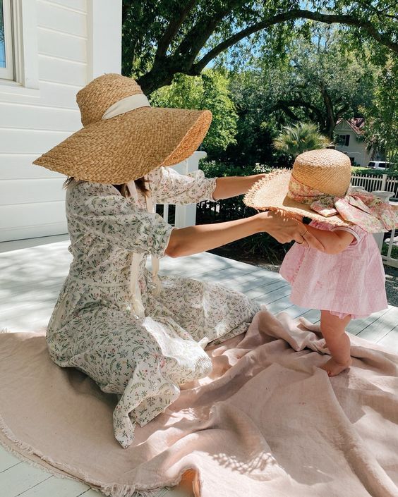 Mother and baby in sun hats enjoy summer on porch, surrounded by nature and bright sunlight.