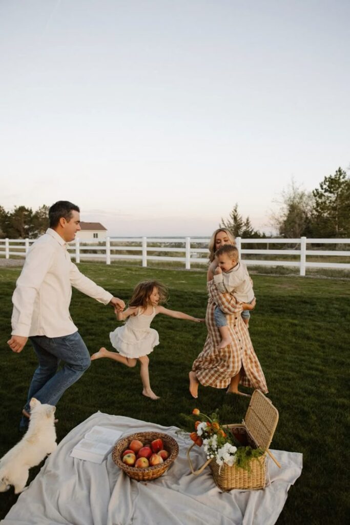 Family enjoying a picnic outdoors with children playing on grassy field near a basket of apples and flowers.