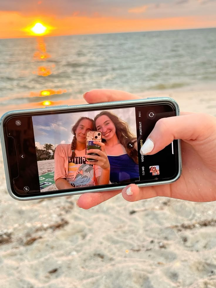 Phone capturing friends' selfie on a beach during sunset, featuring a vibrant orange sky and calm waves.