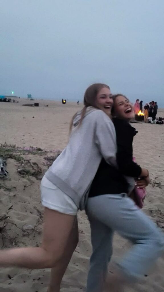 Two friends laughing and hugging on a beach at dusk, with a group gathered around a fire in the background.