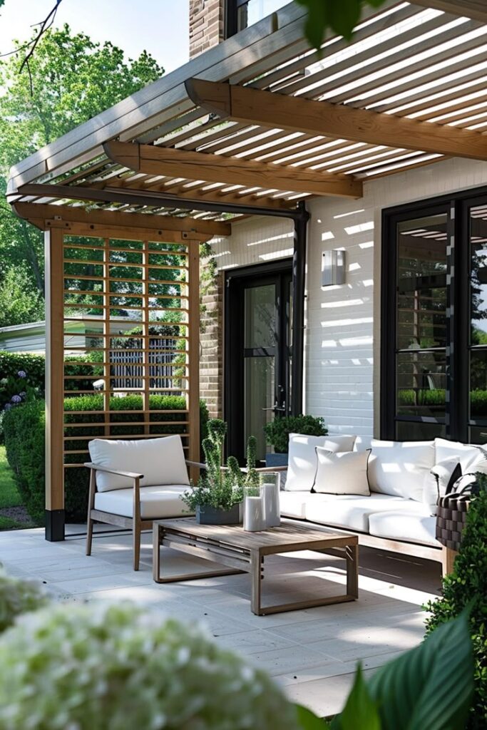 Modern patio with white sofas under a wooden pergola. Lush greenery surrounds the relaxing outdoor space.