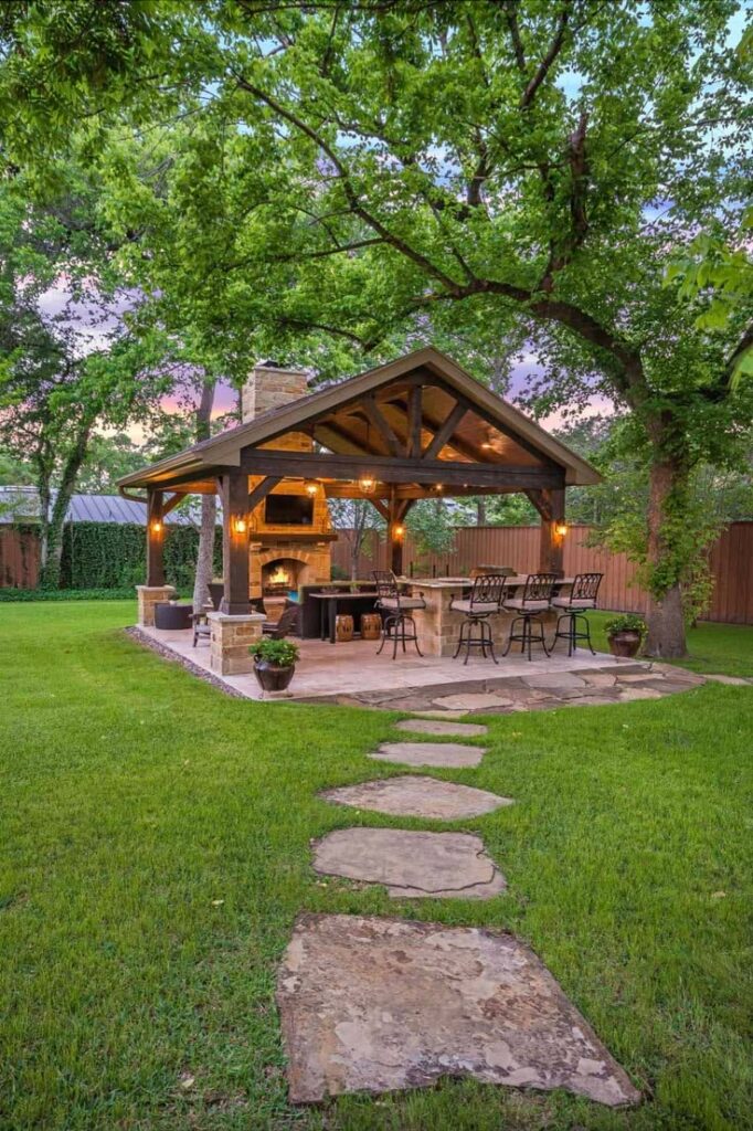 Charming backyard patio with pergola, outdoor kitchen, and bar seating, surrounded by lush green trees and lawn.