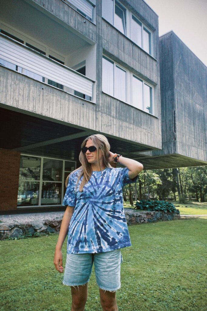 Woman in tie-dye shirt and sunglasses standing on lush lawn near modern building on a sunny day.