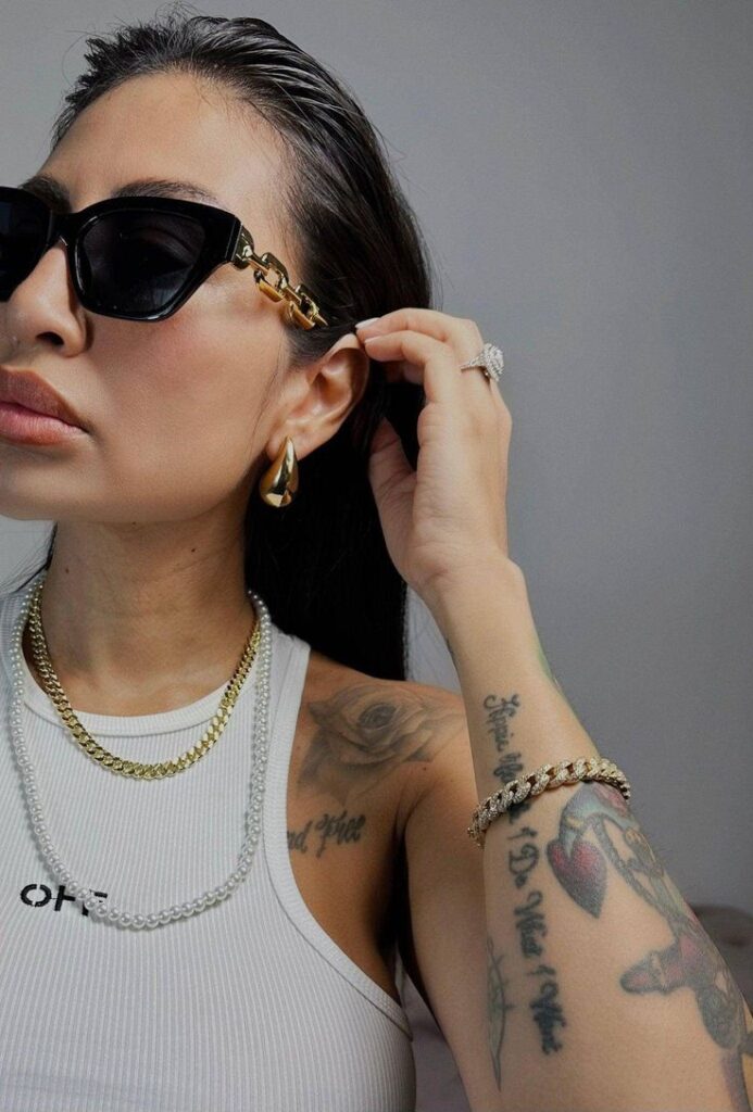 Stylish woman in sunglasses, white top, layered necklaces, and tattoos adjusting hair for a chic look.