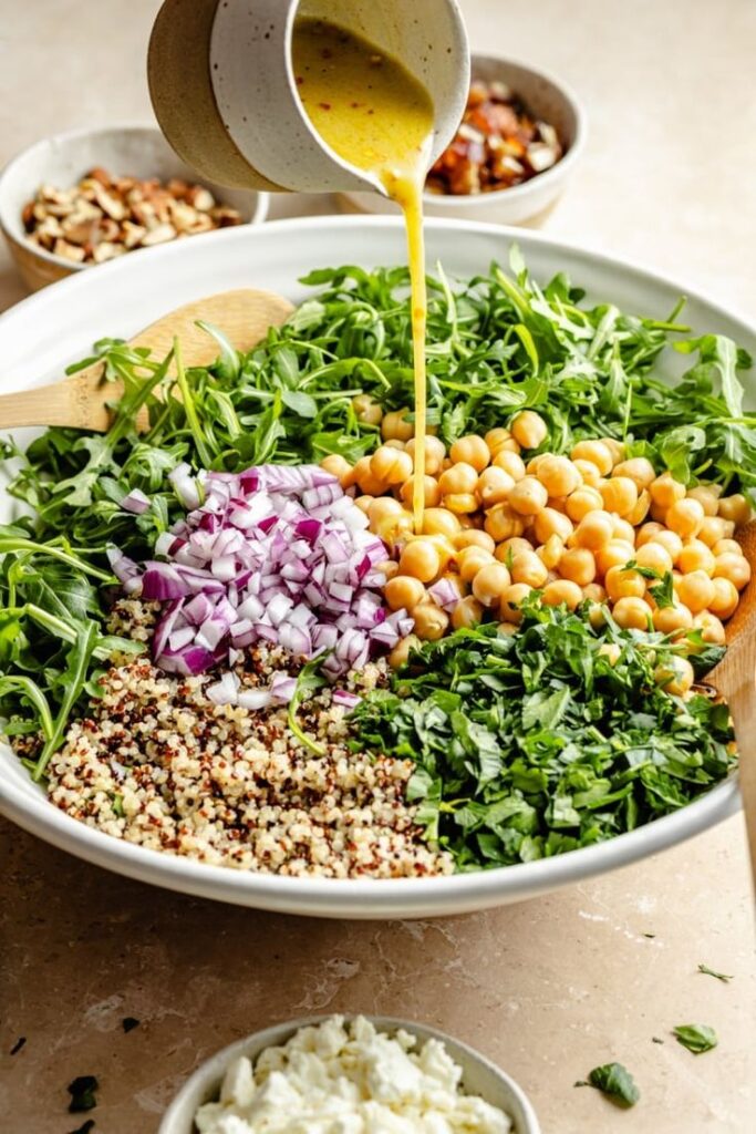 Fresh quinoa salad with chickpeas, arugula, red onion, and dressing being poured in a large white bowl.