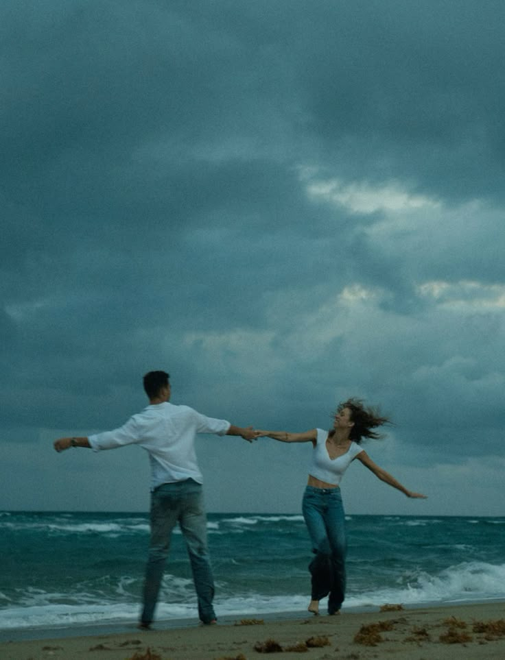 Couple dancing on a moody beach under cloudy skies, capturing a carefree and romantic moment by the ocean.