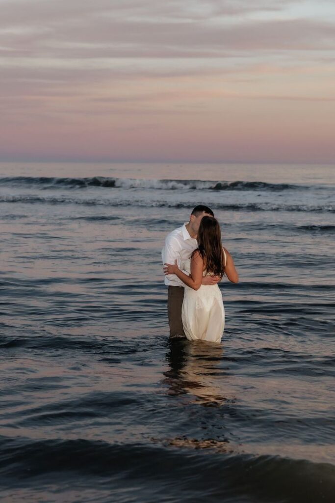 Couple kissing in the ocean during a beautiful sunset, surrounded by gentle waves and colorful sky.