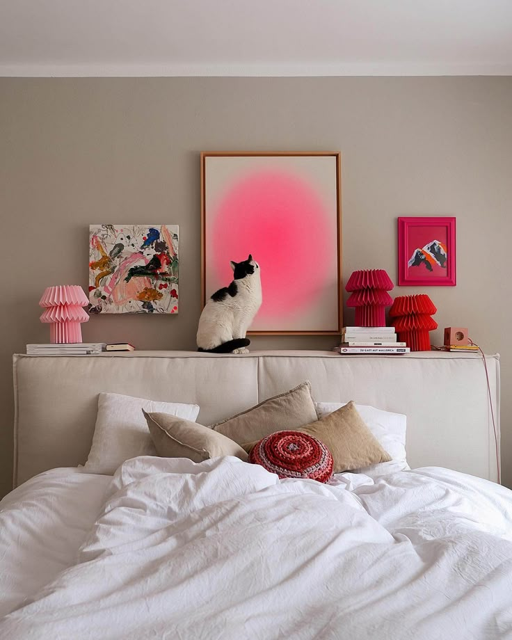 Cozy bedroom scene with a cat on headboard, colorful art, pink lamps, and cushions.