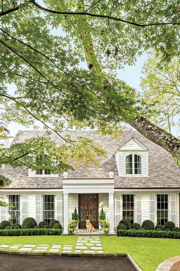 Charming white cottage with a gabled roof, surrounded by lush greenery and a pathway, featuring a dog at the entrance.