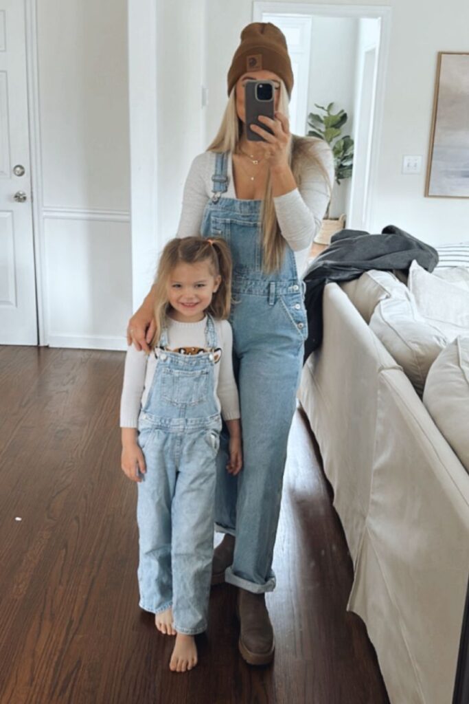 Mother and daughter in matching denim overalls take a mirror selfie in a cozy living room.