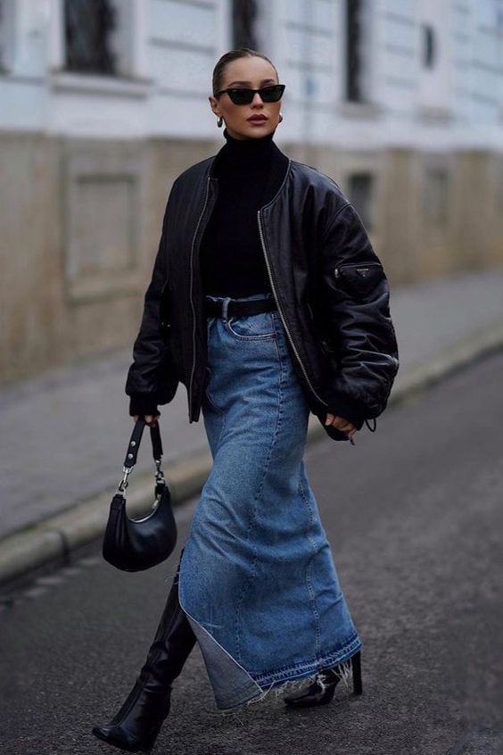 Fashion-forward woman in black jacket, denim skirt, boots, and sunglasses walking on city street, carrying a handbag.