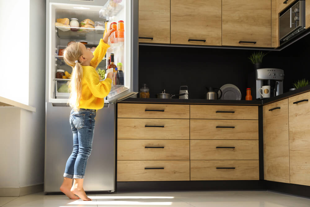 Child in a bright kitchen reaching into a refrigerator, wearing a yellow sweater and jeans.