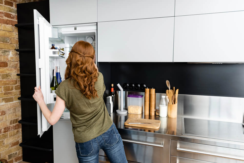 Woman opening fridge in modern kitchen with brick wall and stainless steel countertops.