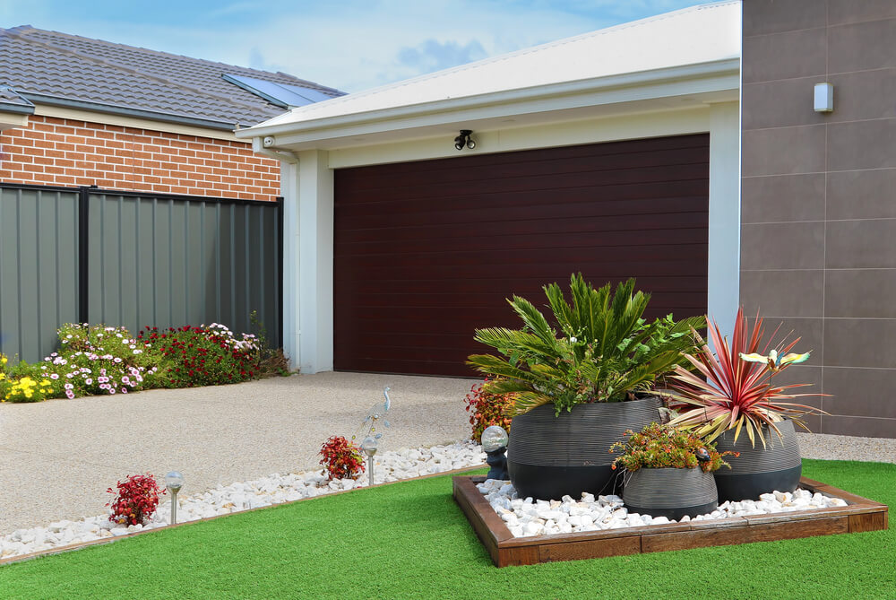 Modern house with landscaped garden and dark garage door, featuring green plants and decorative gravel.