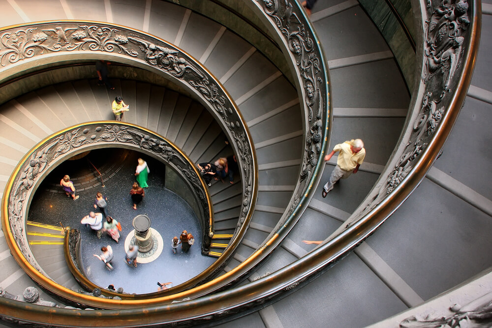 Spiral staircase with ornate railing and people walking, viewed from above in a museum setting.