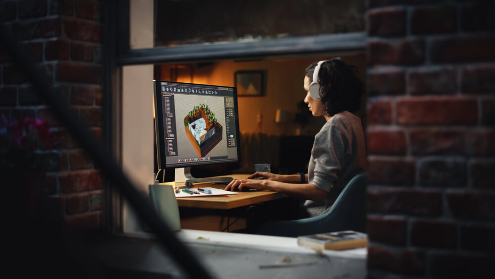 Person wearing headphones working on 3D design on a computer in a cozy room with brick walls.