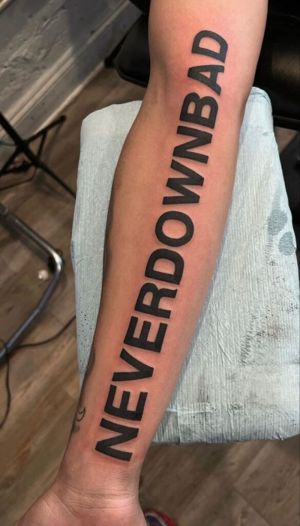 Forearm tattoo with NEVER DOWN BAD in bold black lettering.