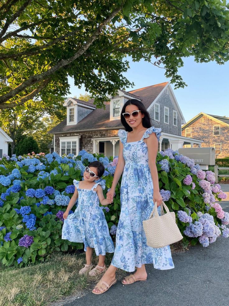 Mother and daughter in matching floral dresses, standing by hydrangeas, with a charming cottage background.