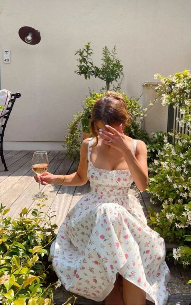 Woman in floral dress sitting on patio with wine, surrounded by greenery and sunlight.