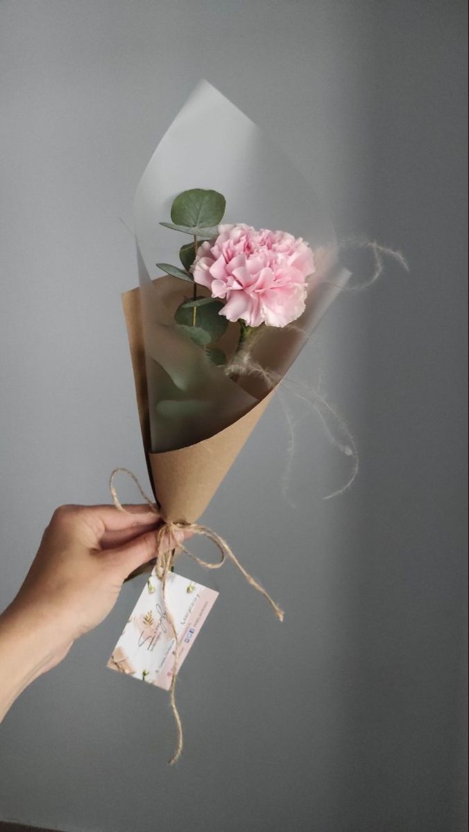 Hand holding a bouquet with a pink carnation and eucalyptus, wrapped in brown paper with a gift tag.