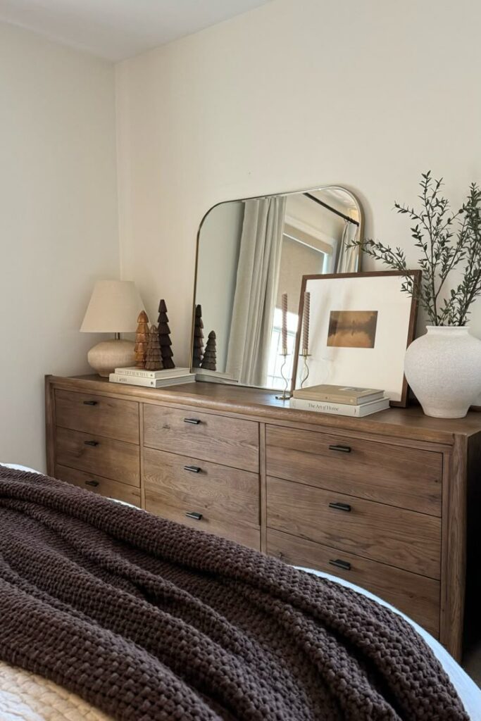 Cozy bedroom with wooden dresser, mirror, and decor items including books, candles, and a vase with greenery.