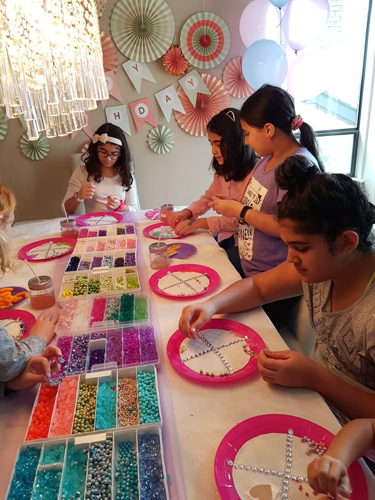 Children at a birthday party making crafts with colorful beads and rhinestones on a decorated table.