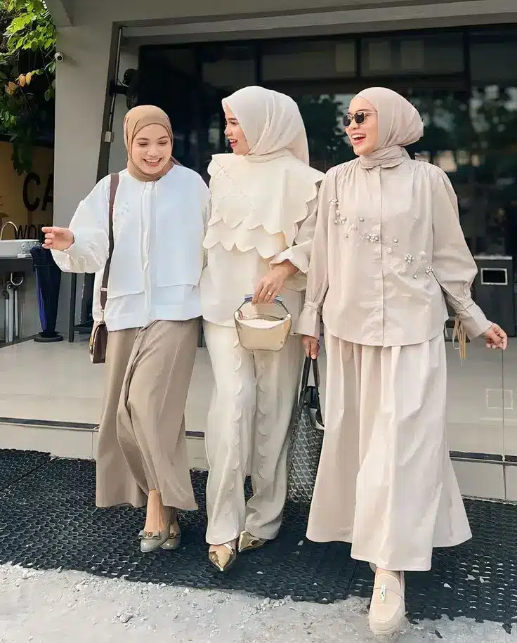 Three women in stylish beige and white outfits with hijabs, walking happily together outside a building.