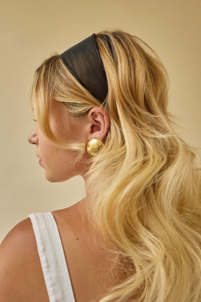 Blonde woman with wavy hair wearing large gold earrings and a black hairband, side profile shot.