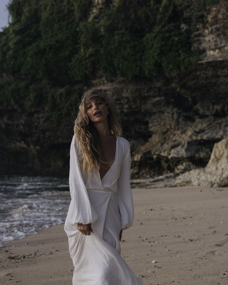 Woman in white dress walking along a secluded beach with rocky cliffs in the background.