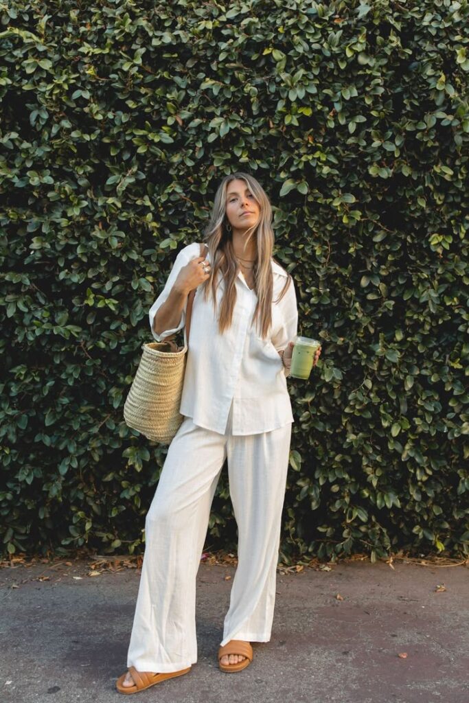 Woman in white outfit with straw bag, holding drink, stands against green leafy background.