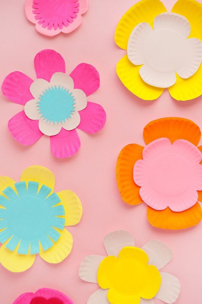 Colorful paper plate flowers on a pink background, featuring bright shades of pink, yellow, and blue petals.