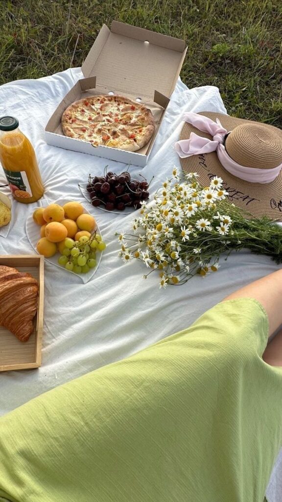 Picnic with pizza, fresh fruits, juice, and daisies on a white blanket in a grassy field. Summer outdoor scene.