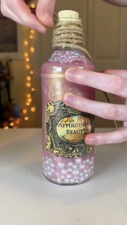 Hand decorating a pink vintage bottle labeled Aphrodite's Beauty with pearls and twine, blurred lights in background.